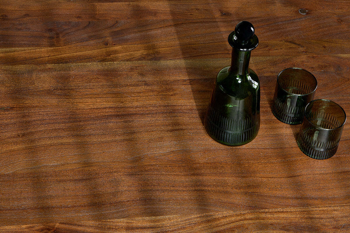 The Anbu Acacia Dining Table in Washed Walnut by nkuku features a green glass decanter with a stopper and two matching tumblers. Diagonal shadows from window blinds fall across the wood, with the set arranged in the upper right of the image.
