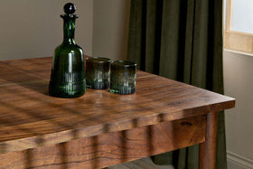 A dark green glass decanter with a round stopper and two matching tumblers sit on the nkuku Anbu Acacia Dining Table in Washed Walnut, sunlight casting soft shadows. Behind the table, a green curtain frames a sunlit window.