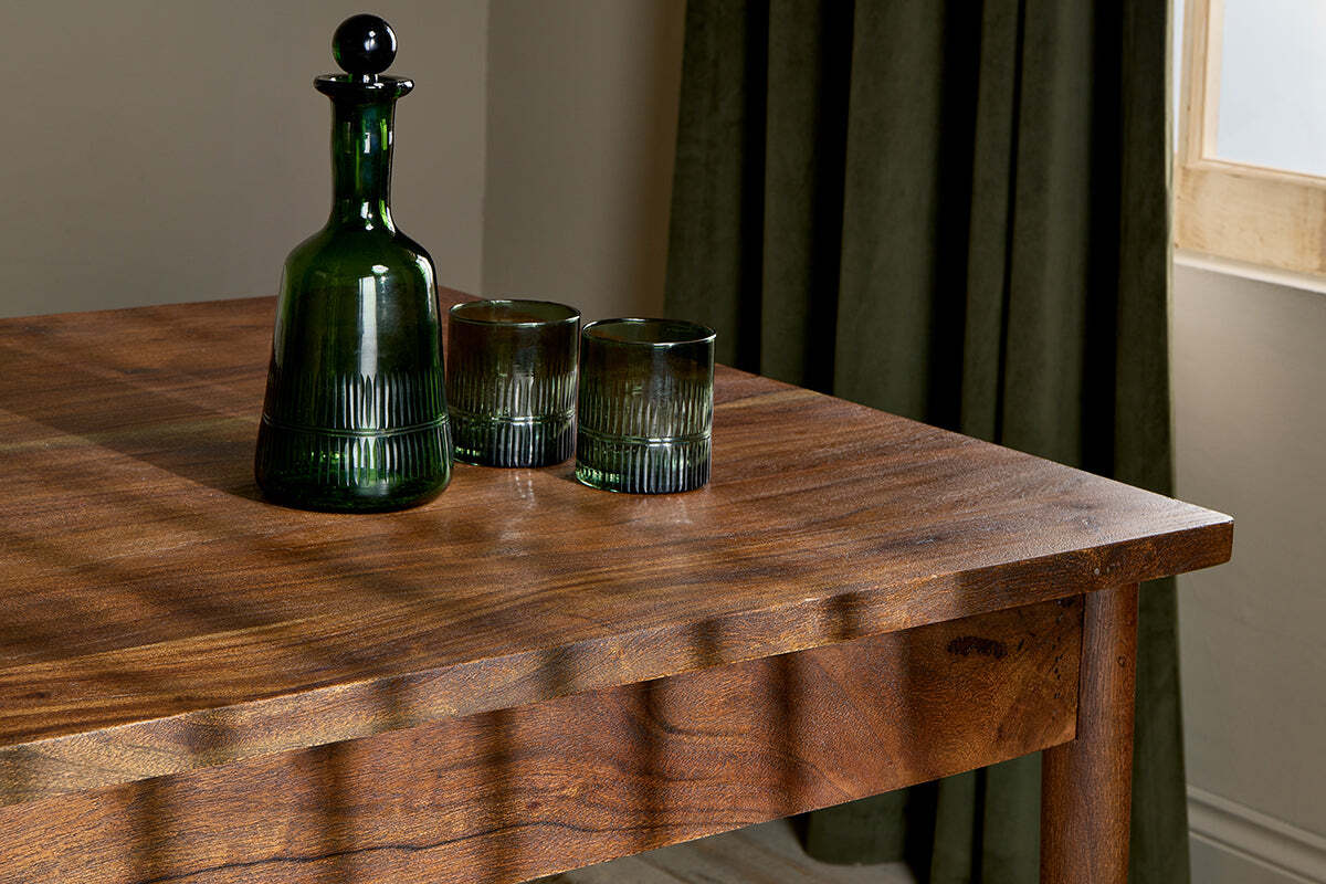 A dark green glass decanter with a round stopper and two matching tumblers sit on the nkuku Anbu Acacia Dining Table in Washed Walnut, sunlight casting soft shadows. Behind the table, a green curtain frames a sunlit window.