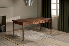 The Anbu Acacia Dining Table by nkuku, finished in washed walnut with tapered legs, stands on a textured beige rug in a sunlit room featuring wooden floors, olive green curtains, modern chandelier, and neutral minimalist walls.