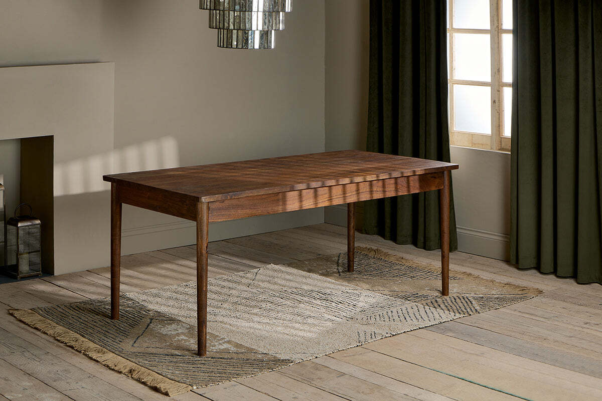 The Anbu Acacia Dining Table by nkuku, finished in washed walnut with tapered legs, stands on a textured beige rug in a sunlit room featuring wooden floors, olive green curtains, modern chandelier, and neutral minimalist walls.
