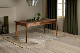 The nkuku Anbu Acacia Dining Table in Washed Walnut sits on a patterned rug, topped with three green glasses, a green bottle, and a vase of flowers. Behind it, dark green curtains frame the window.