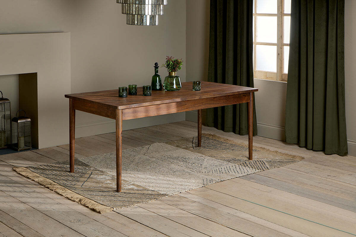 The nkuku Anbu Acacia Dining Table in Washed Walnut sits on a patterned rug, topped with three green glasses, a green bottle, and a vase of flowers. Behind it, dark green curtains frame the window.