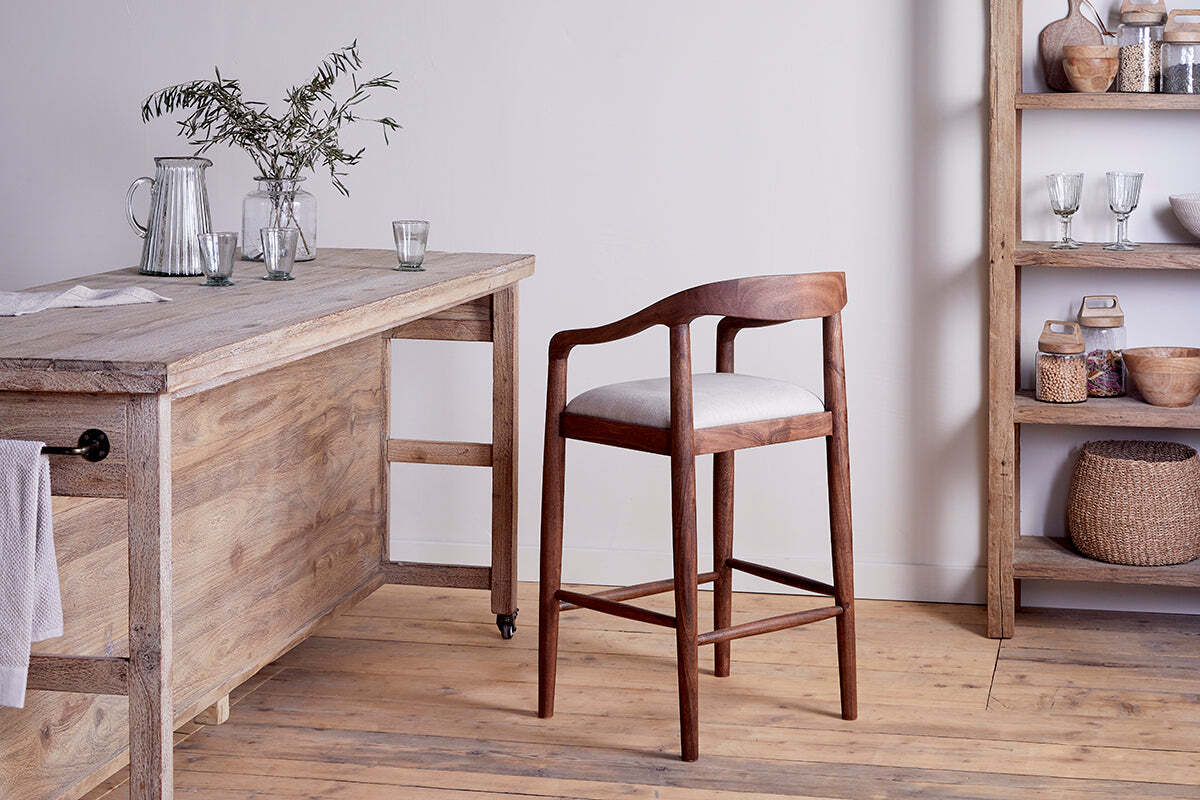 The nkuku Anbu Acacia Upholstered Counter Chair in Washed Walnut, featuring a curved back and light cushion, sits by a rustic acacia table with glassware and foliage, creating a cozy, minimalist space.