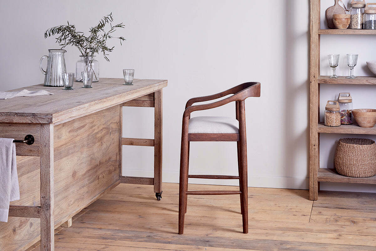 An Anbu Acacia Upholstered Counter Chair by nkuku in washed walnut, with a curved backrest, sits beside a rustic wood table adorned with glassware and greenery. An open wooden shelf holds bowls and jars; acacia floors and neutral walls complete the room.