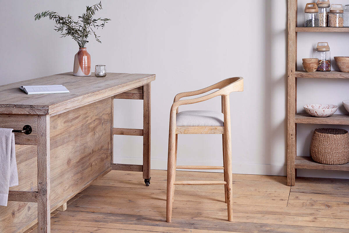 A light wooden desk with a book, candle, and vase stands on a wooden floor. In front is the nkuku Anbu Mango Wood Upholstered Counter Chair - Natural. To the right, shelves hold jars and baskets against a white wall.