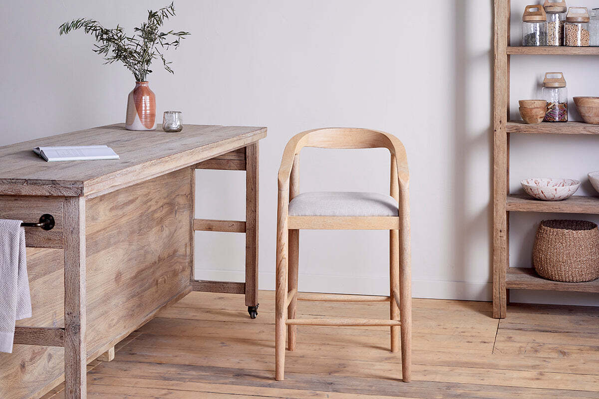 The Anbu Mango Wood Upholstered Counter Chair – Natural by nkuku stands beside a rustic table. A ceramic vase with greenery, a glass, and shelves with jars and baskets complete the minimalist wooden-floored room.