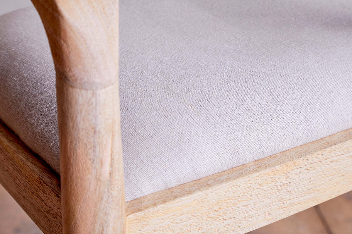 Close-up of the nkuku Anbu Mango Wood Upholstered Counter Chair - Natural, featuring a textured light beige cushion, smooth rounded armrests, and a minimalist frame highlighting the natural wood grain and light finish.
