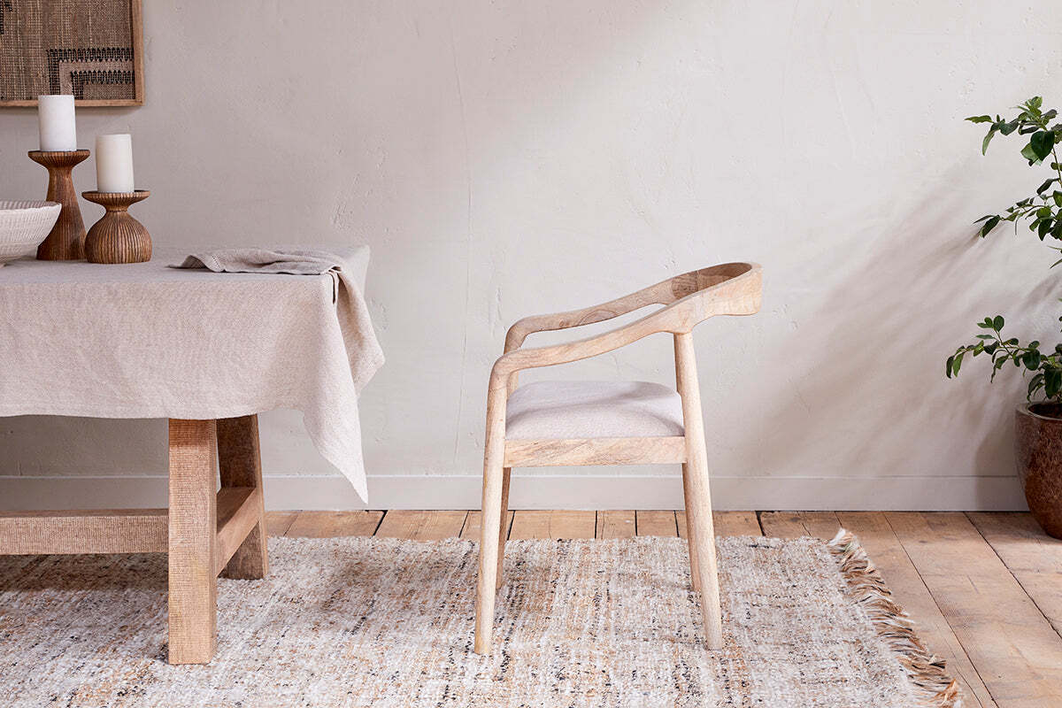 The Anbu Mango Wood Upholstered Dining Chair by nkuku sits beside a matching table with a beige cloth, wooden candle holders, and candles. A woven rug and potted plant add to the calm, minimalist room.