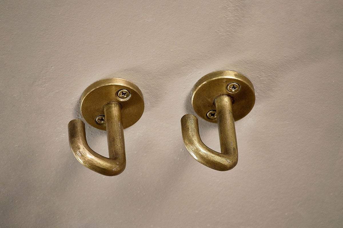 The Aniko Iron Ceiling Hook - Brass (Set of 2) by nkuku features brass-finished hooks with rounded bases and curved arms, perfect for organizing kitchen storage or hanging items, and mounted side by side on a light-colored wall.