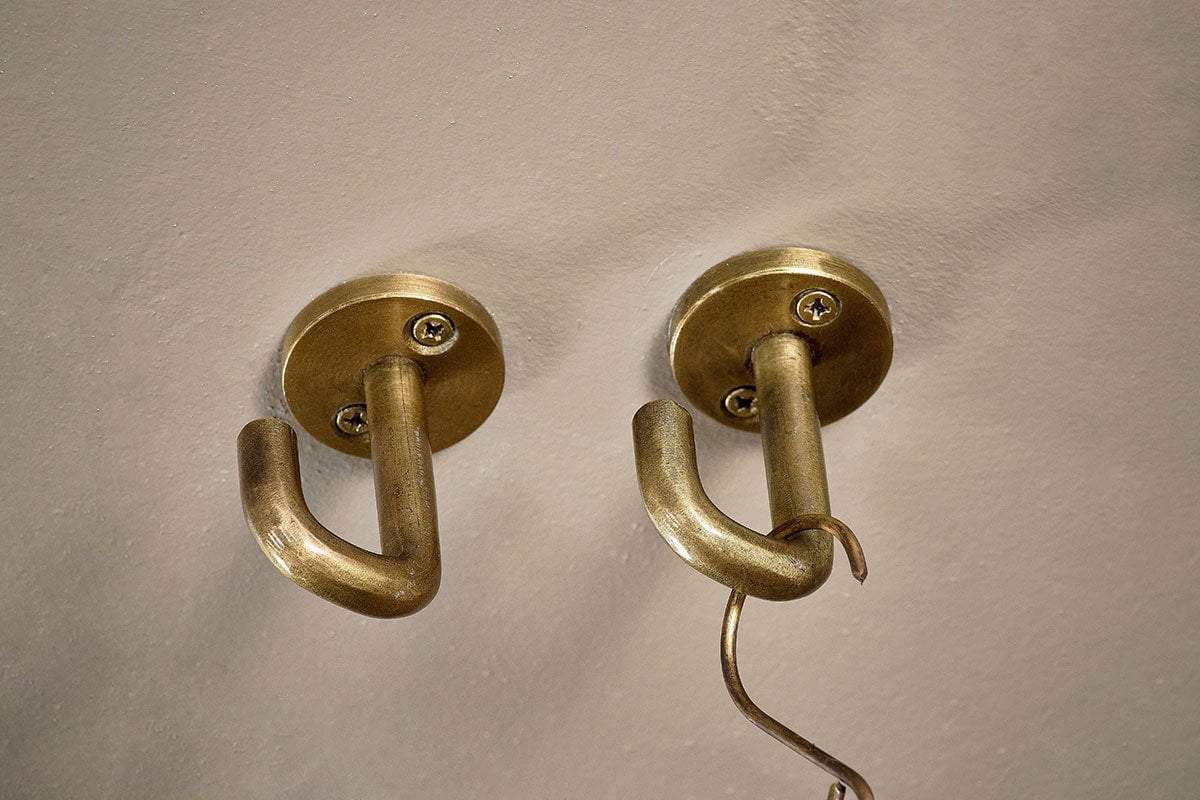 The nkuku Aniko Iron Ceiling Hook - Brass (Set of 2) is mounted side by side on a beige wall. Each hook features a round base with two visible screws. A thin metal object hangs from the right hook, while the left one remains empty.