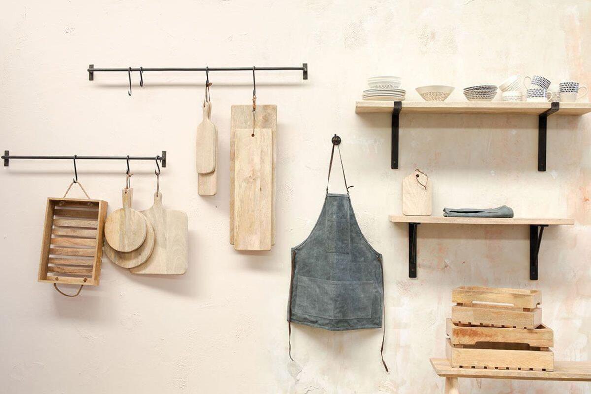 A minimalist kitchen wall features two black metal racks with wooden cutting boards, utensils, and a gray apron on nkuku’s Aniko Iron Hook (Set of 2). Wooden shelves hold stacked plates and bowls; crates are arranged neatly below.