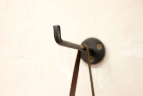 A close-up of a black, hand-cast Aniko Iron Hook by nkuku (Set of 2), mounted with two screws on a beige wall, displays a brown strap hanging from it; another L-shaped hook is slightly out of focus in the background.