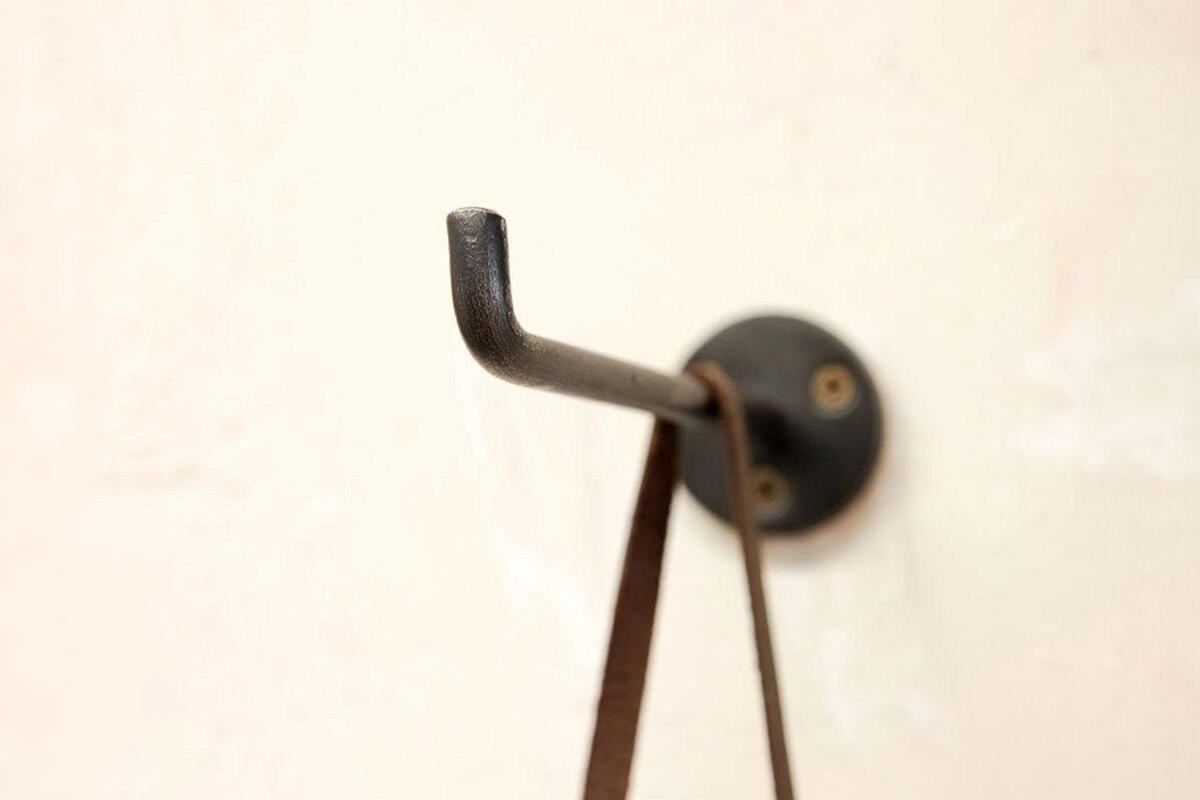A close-up of a black, hand-cast Aniko Iron Hook by nkuku (Set of 2), mounted with two screws on a beige wall, displays a brown strap hanging from it; another L-shaped hook is slightly out of focus in the background.