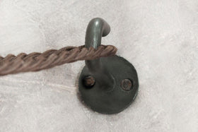 A close-up of an nkuku Aniko Iron Hook (Set of 2), wall-mounted with two screws on a light gray textured surface. A brown braided cord hangs over the hook, showcasing its sturdy iron build and minimalist design.