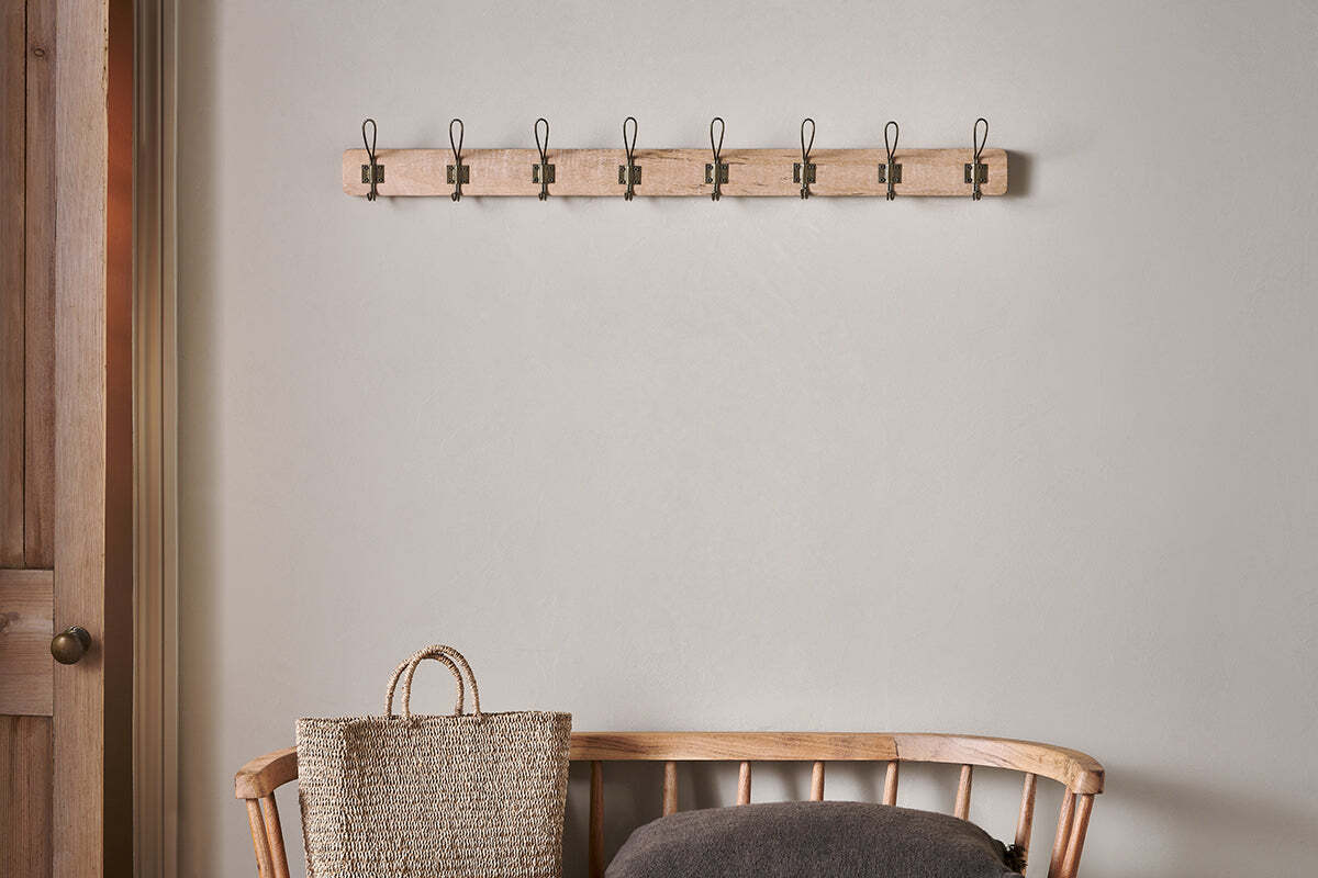 The nkuku Ankita Mango Wood & Iron Hooks - Natural & Antique Brass - Large is mounted on a beige wall above a wooden bench. On the bench are a woven handled basket and dark gray cushion; a closed wooden door sits to the left in this serene setting.
