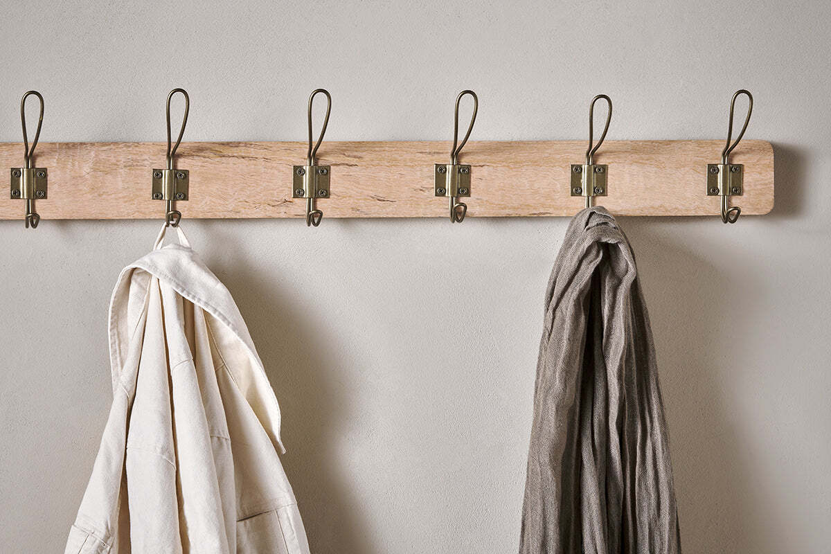 The Ankita Mango Wood & Iron Hooks rack by nkuku, featuring five antique brass hooks on natural wood, is mounted on a light wall. A white garment hangs on the leftmost hook; a gray one is second from the right. The other hooks are empty.