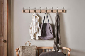 The Ankita Mango Wood & Iron Hooks by nkuku in natural and antique brass hold a cream jacket, grey tote bag, and grey scarf. Below, a woven tote rests on a wooden bench with a dark grey cushion.