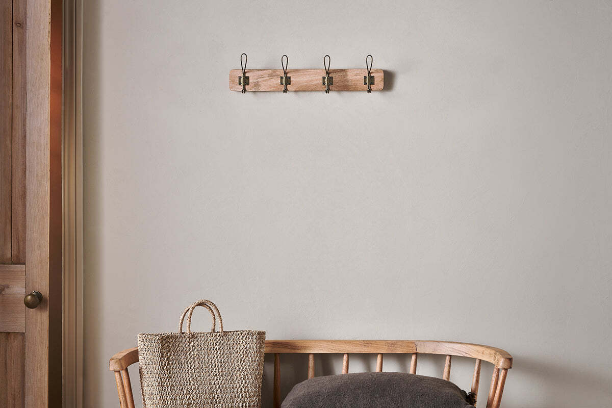 A wooden bench with a dark cushion and woven straw bag sits against a light beige wall. Above, the nkuku Ankita Mango Wood & Iron Hooks in Natural & Antique Brass (Small) are mounted. A wooden door is partially visible on the left.