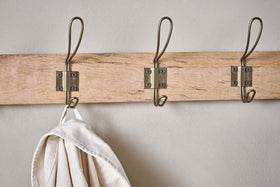The Nkuku Ankita Mango Wood & Iron Hooks - Natural & Antique Brass - Small features three artisan metal hooks mounted on a wooden rack. The left hook holds a cream-colored cloth or bag, while the other two are empty and attached with visible screws.