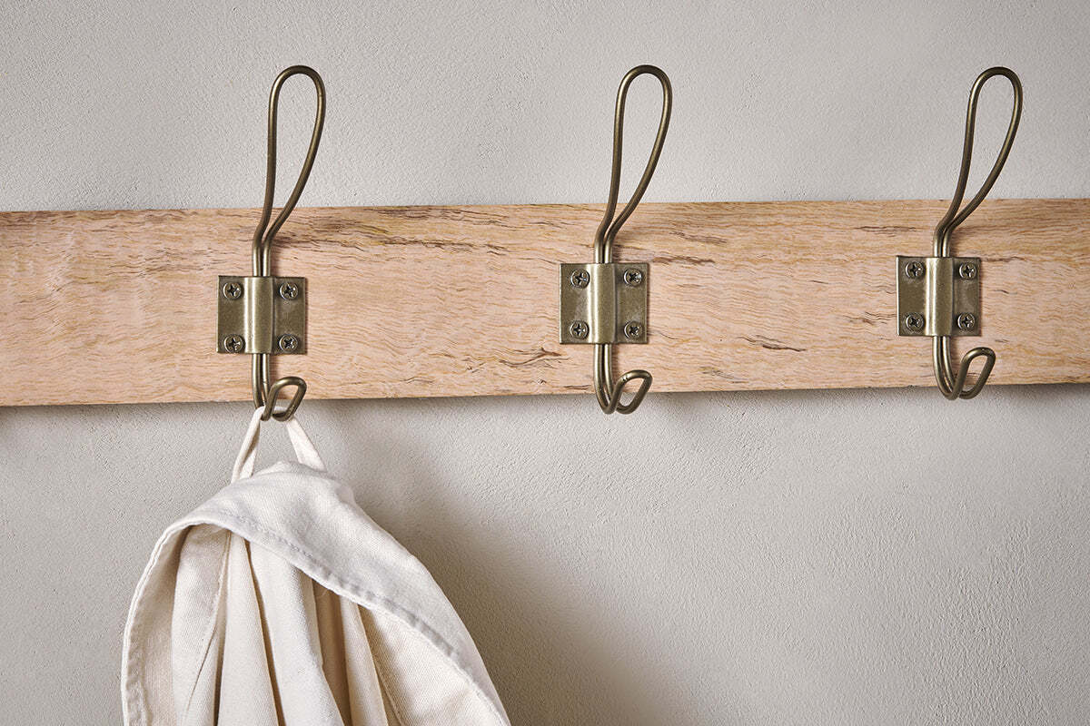 The Nkuku Ankita Mango Wood & Iron Hooks - Natural & Antique Brass - Small features three artisan metal hooks mounted on a wooden rack. The left hook holds a cream-colored cloth or bag, while the other two are empty and attached with visible screws.