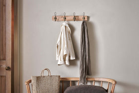 The nkuku Ankita Mango Wood & Iron Hooks in Natural & Antique Brass (Small) are mounted above a wooden bench. A light shirt and gray scarf hang from the hooks, with a woven bag and dark cushion nearby against a light gray wall.
