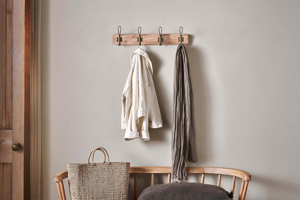 The nkuku Ankita Mango Wood & Iron Hooks in Natural & Antique Brass (Small) are mounted above a wooden bench. A light shirt and gray scarf hang from the hooks, with a woven bag and dark cushion nearby against a light gray wall.