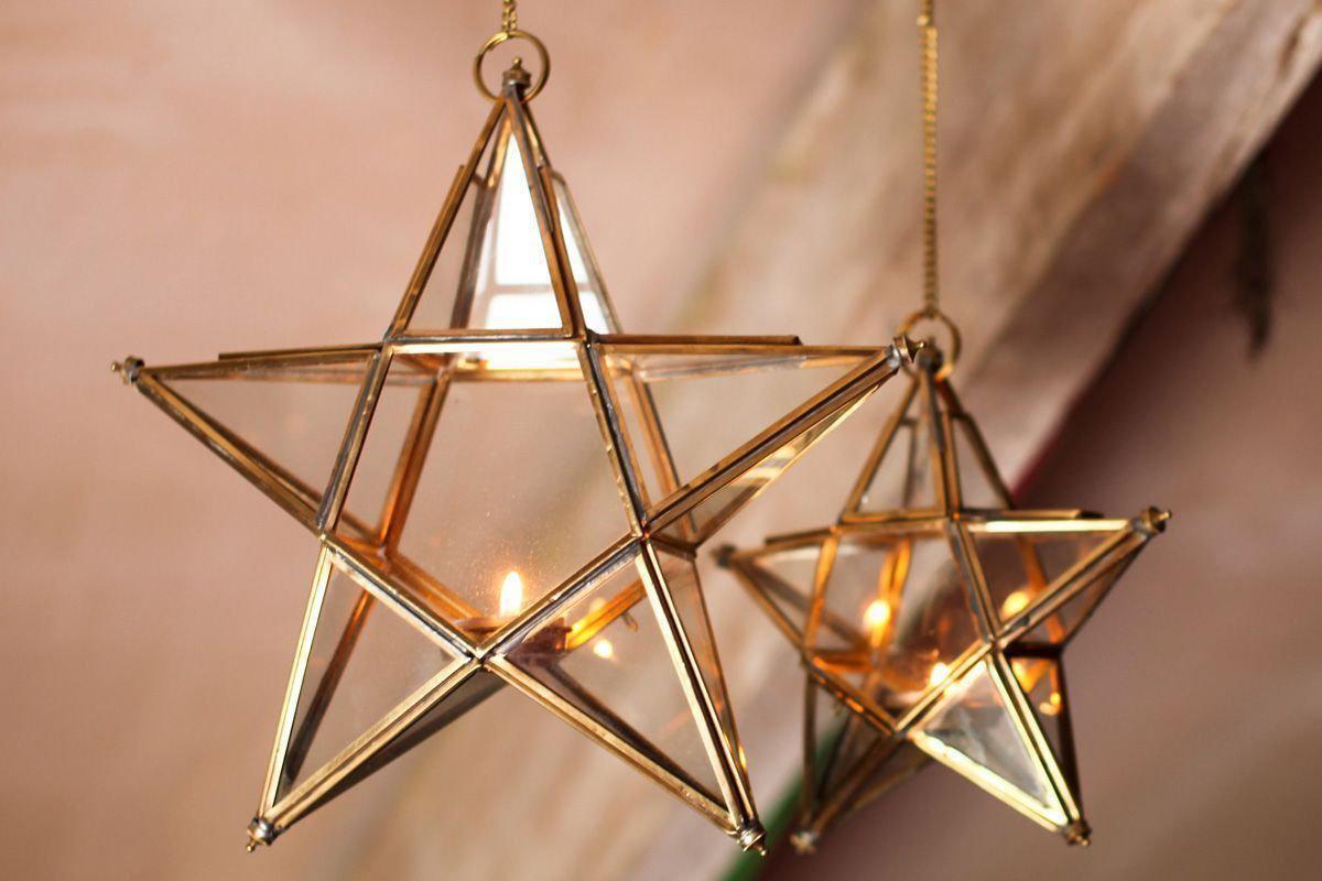 Two nkuku Antique Brass Glass Star t-light holders hang side by side, each with a glowing candle. Their gold frames and handmade glass catch the warm, blurred beige background light, highlighting the geometric lanterns.