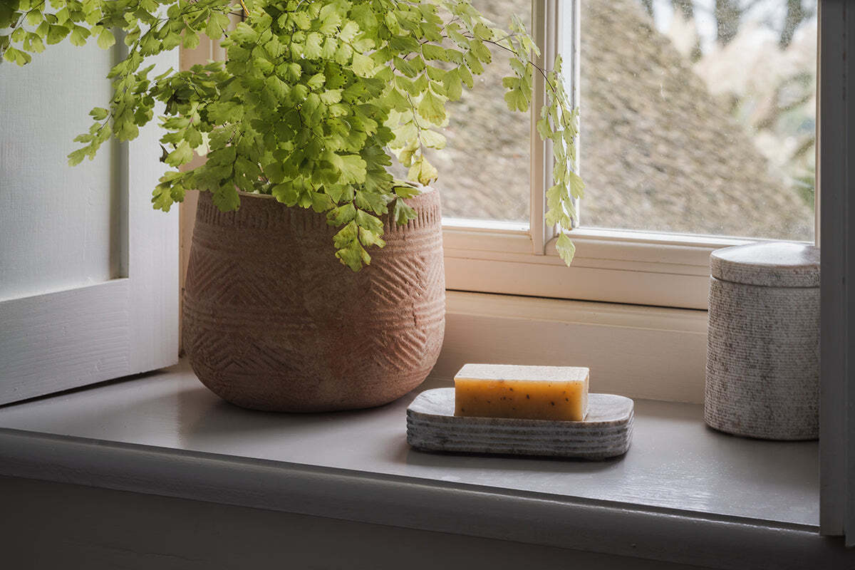 A leafy fern in a textured clay pot sits on a windowsill beside a yellow soap bar resting on the nkuku Asana Marble Soap Dish - Grey, sunlight streaming through the window and highlighting this calm, elegant scene.