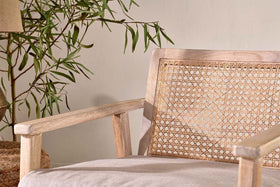 The nkuku Atri Mango Wood & Cane Occasional Chair features sustainable mango wood, a woven backrest, and a light beige cushion. Beside it, a leafy plant and woven basket enhance the tranquil, natural setting against a neutral wall.