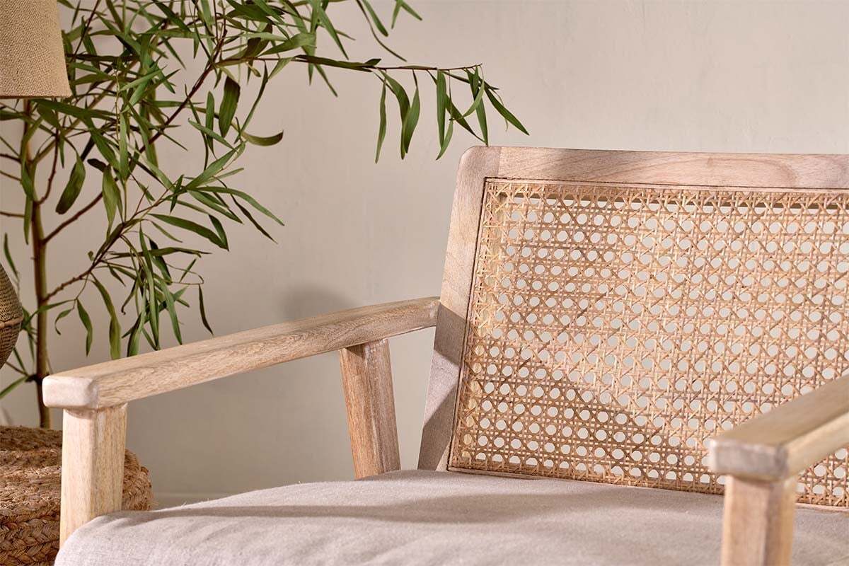 The nkuku Atri Mango Wood & Cane Occasional Chair features sustainable mango wood, a woven backrest, and a light beige cushion. Beside it, a leafy plant and woven basket enhance the tranquil, natural setting against a neutral wall.