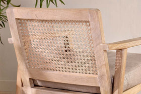 Close-up of the nkuku Atri Mango Wood & Cane Occasional Chair featuring a woven backrest, light natural wood finish, and cushioned seat. A green leafy plant appears in the upper left corner against a beige wall backdrop.