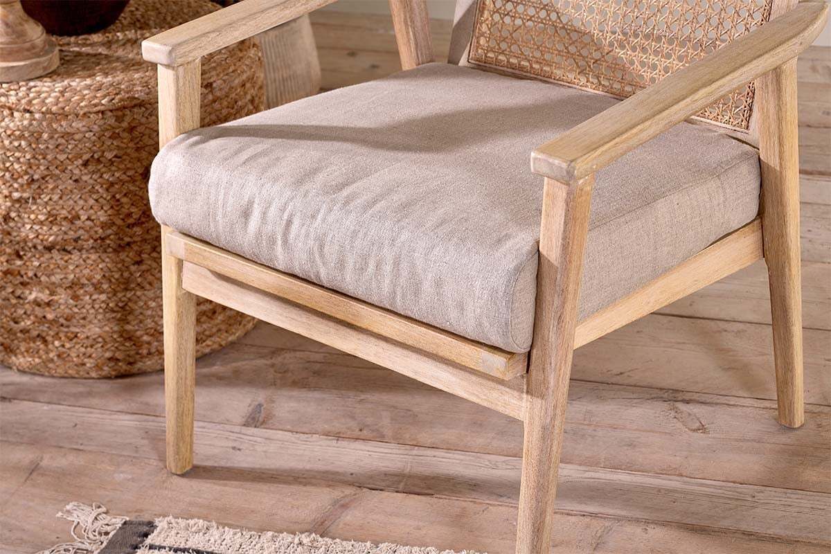 The nkuku Atri Mango Wood & Cane Occasional Chair features a beige cushioned seat and wicker backrest on a wooden floor, with a woven basket nearby and a fringed rug visible in the corner.