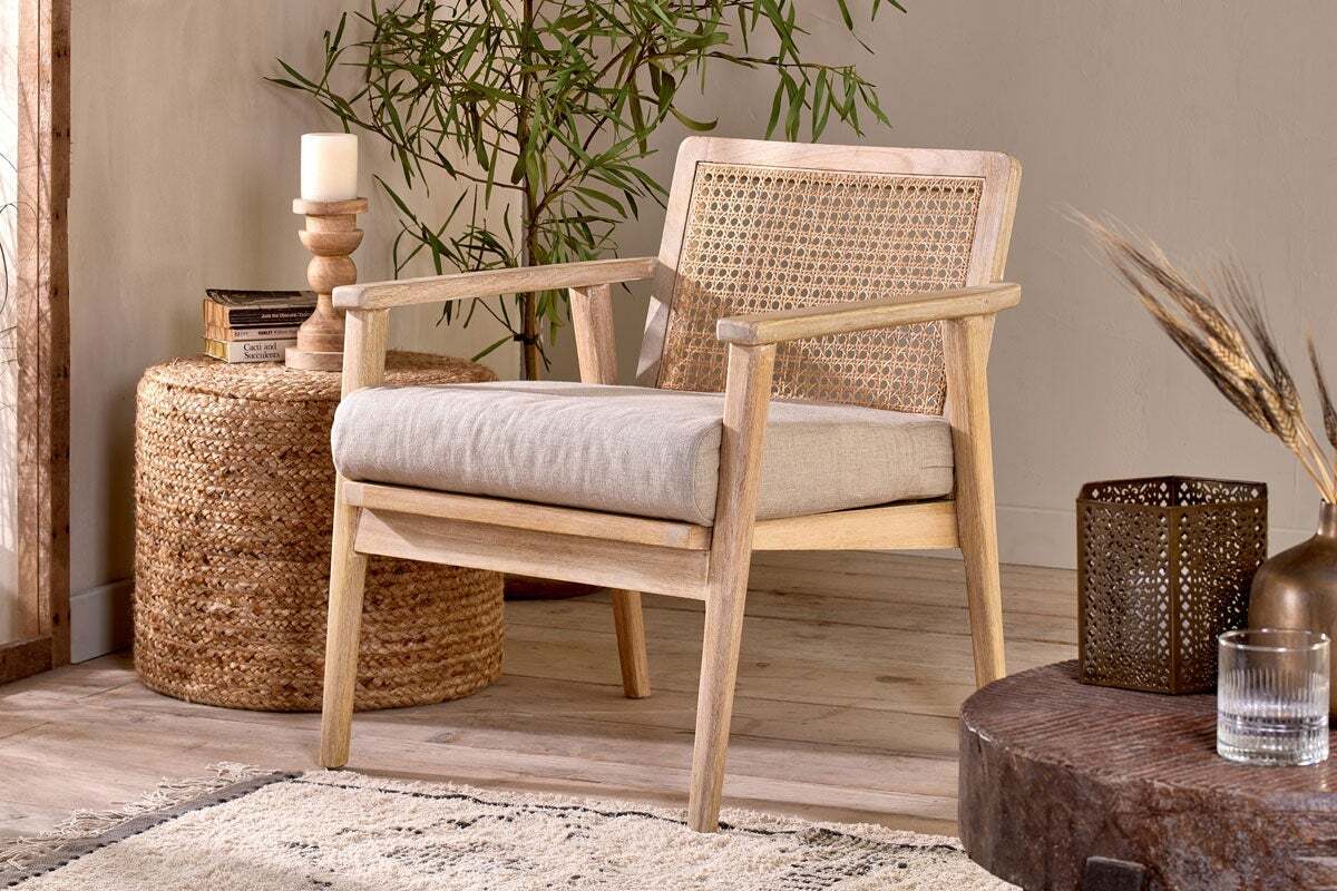 The nkuku Atri Mango Wood & Cane Occasional Chair, with a woven rattan backrest and beige cushion, rests on a light rug beside a round wicker side table holding a candle and vase, accented by plants for a cozy, natural look.
