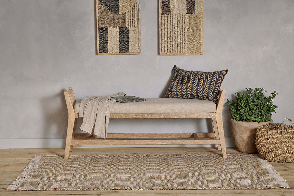 The Avanthi Upholstered Sleigh Bench by nkuku, with natural linen upholstery, sits on a jute rug. Above, woven wall art panels hang. A terracotta potted plant and wicker basket complete the boho-neutral look with chevron pillow and throw.
