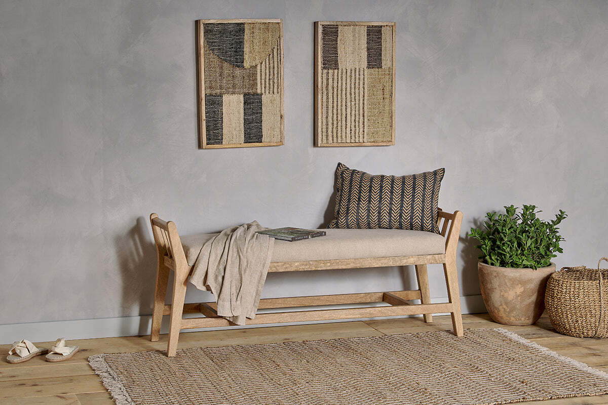 The nkuku Avanthi Upholstered Sleigh Bench in Natural sits against a gray wall with two woven art pieces. It features a patterned pillow on natural linen, a throw and book, with a woven rug in front and a potted plant with basket to the right.