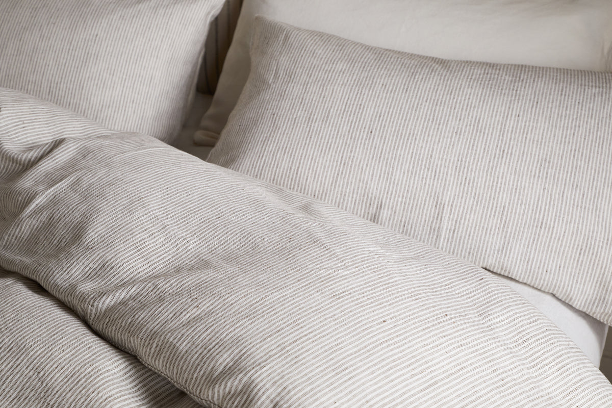 Close-up of a neatly made bed with beige and white pinstriped bedding, featuring the nkuku Borba Hemp & Cotton Duvet Cover in Espresso, along with pillows, creating a cozy and inviting look.