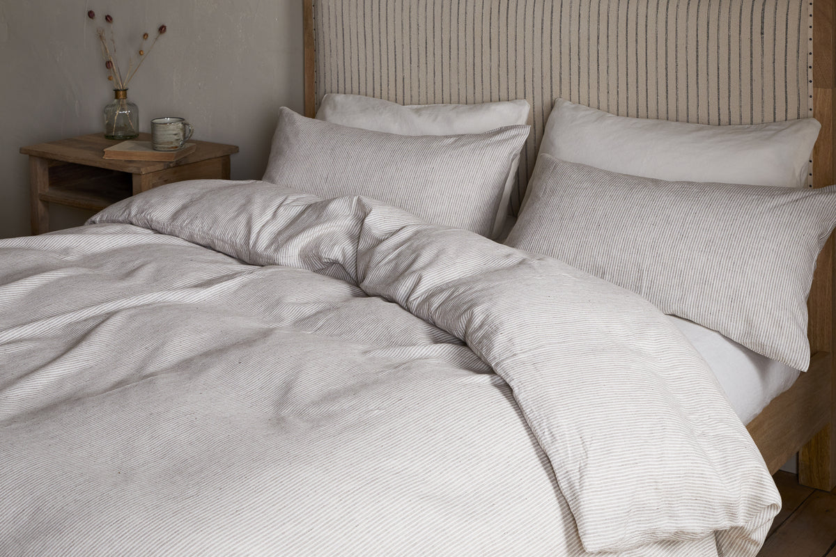 A neatly made bed features the nkuku Borba Hemp & Cotton Duvet Cover in Espresso, with matching pillows, beside a wooden nightstand topped with a clear vase of dried flowers and a small glass jar.