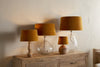 Four Batakha Velvet & Linen Lampshades in Dark Ochre by nkuku create an elegant look on a rustic wooden console, featuring two clear glass bases, one ribbed ceramic base, and one wooden base, all placed against a beige wall.