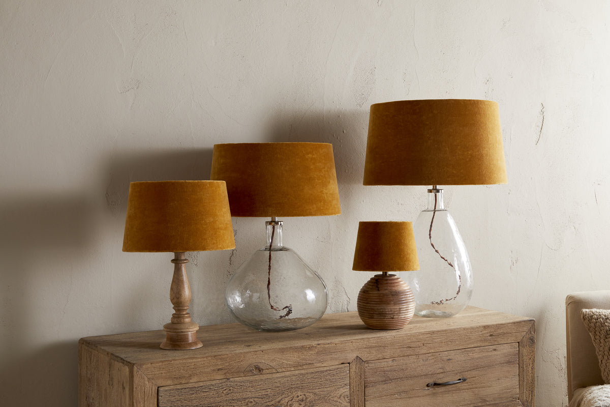 Four table lamps with Batakha Velvet & Linen Lampshades in Dark Ochre by nkuku are arranged on a wooden sideboard; two have clear glass bases, one has a ribbed ceramic base, and one features a turned wooden base, set against a beige wall.