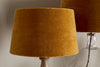 A close-up features the nkuku Batakha Velvet & Linen Lampshade in Dark Ochre, highlighting elegant lighting. A similar lamp with a glass base appears partially visible in the background against a light wall.