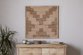 A light wooden chest holds small rustic cups, a tray, and books. Above, the nkuku Bawal Wall Art - Cross - Natural with tan and brown geometric patterns hangs on a white wall. To the left stands a leafy green plant.