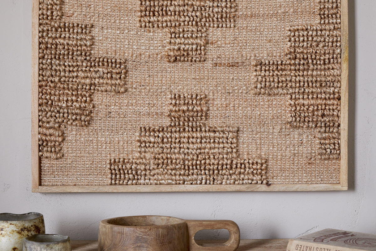 A close-up of the nkuku Bawal Wall Art - Cross - Natural, featuring Indian geometric patterns in a sustainable mango wood frame. Below, on a light wood shelf, are ceramic cups, a handled wooden bowl, and part of a book.