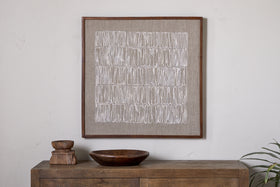 The Birala Hand Stitched Wall Art - Natural by nkuku, featuring white linear patterns inspired by Indian Brahmic script, is framed in wood and displayed above a rustic, sustainable mango wood sideboard adorned with decor and greenery.