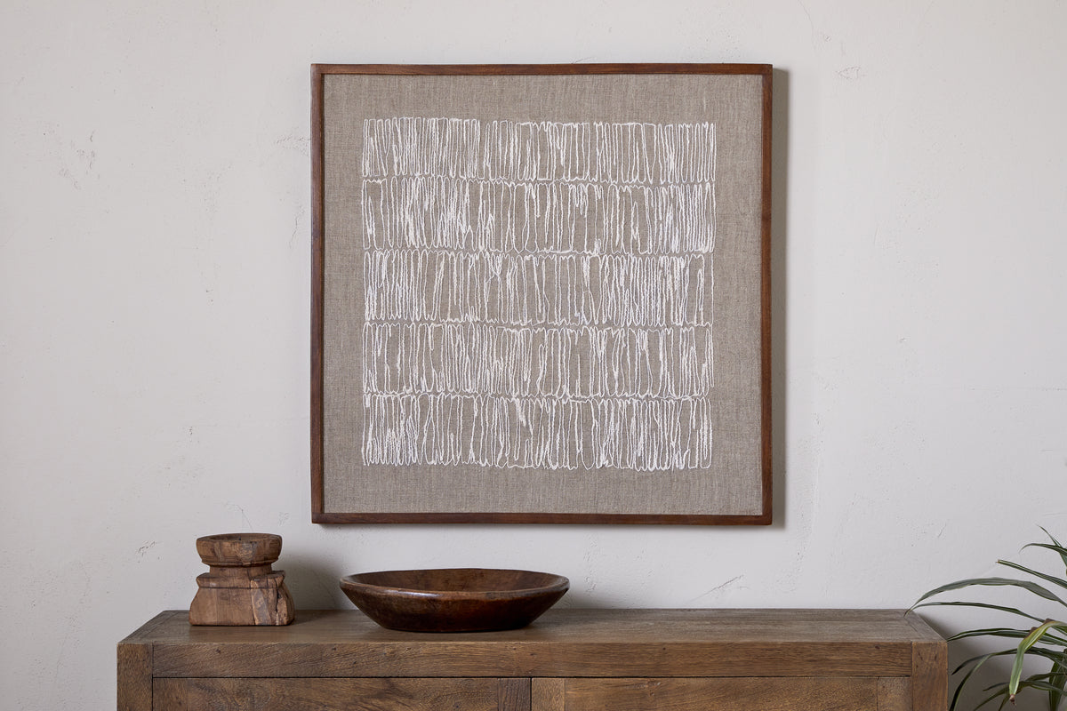 The Birala Hand Stitched Wall Art - Natural by nkuku, featuring white linear patterns inspired by Indian Brahmic script, is framed in wood and displayed above a rustic, sustainable mango wood sideboard adorned with decor and greenery.