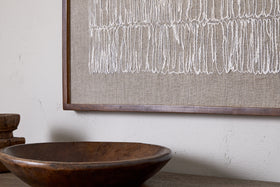 A rustic mango wood bowl sits on a wooden surface in front of the nkuku Birala Hand Stitched Wall Art - Natural, which features vertical white lines on textured beige fabric; stacked wooden objects are partially visible to the left.