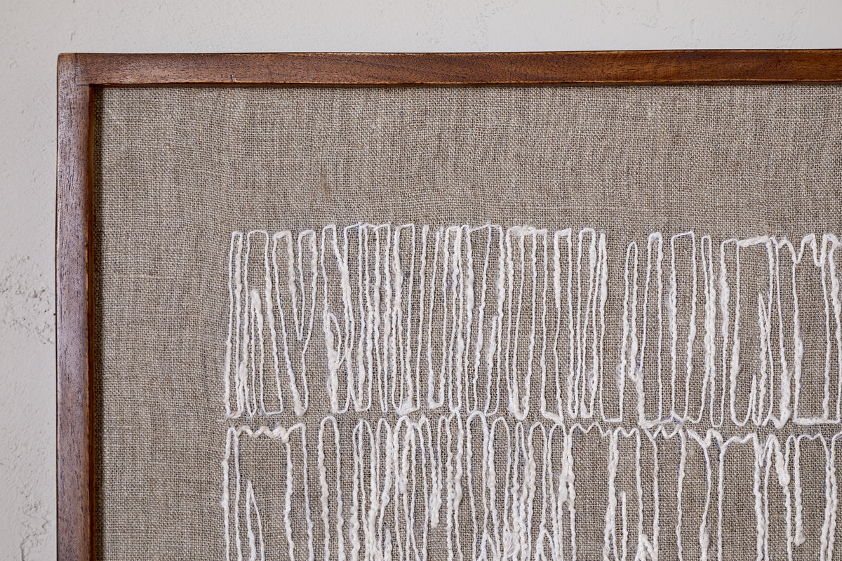 The Birala Hand Stitched Wall Art - Natural by nkuku displays irregular vertical white threads on beige fabric, framed in sustainable mango wood, highlighting the textured abstract pattern against a softly textured wall.