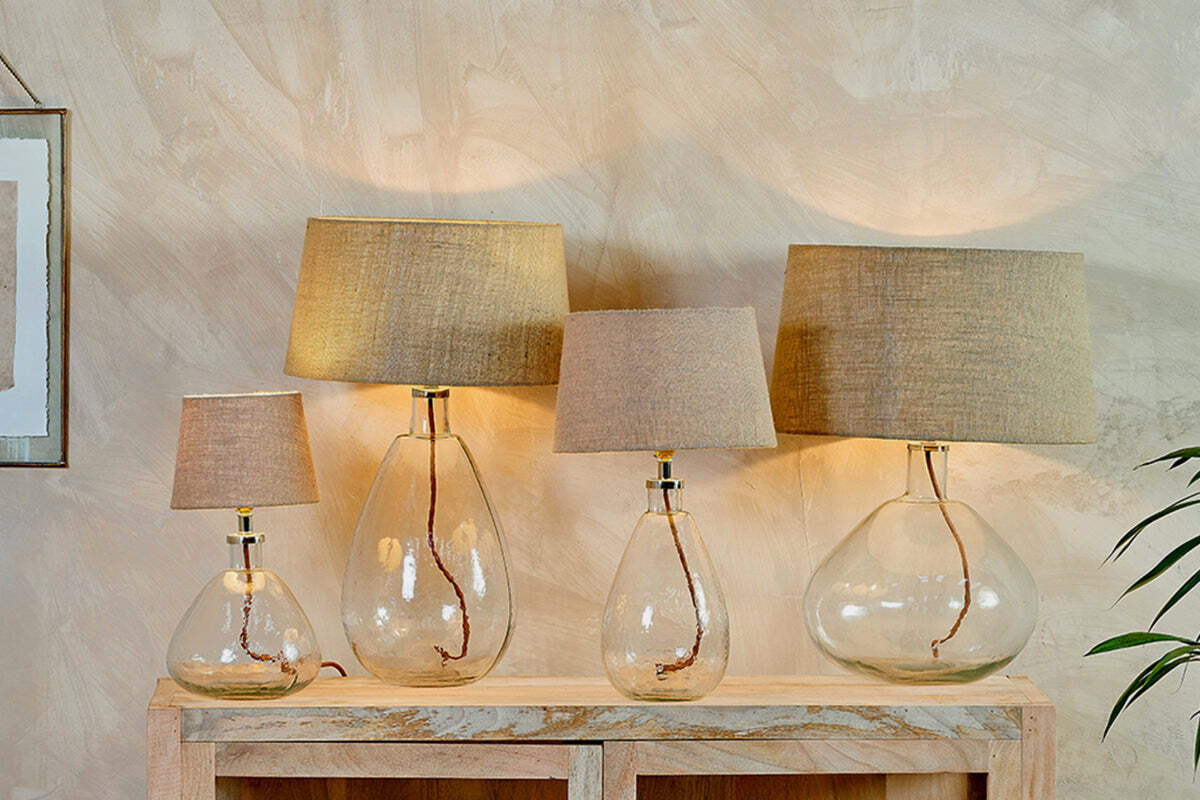 Four nkuku Baba Recycled Glass Lamps in Clear, Large Wide, with beige fabric shades, are arranged on a wooden console. Their recycled glass bases reveal twisted brown cords, casting a soft, warm glow against a textured beige wall.