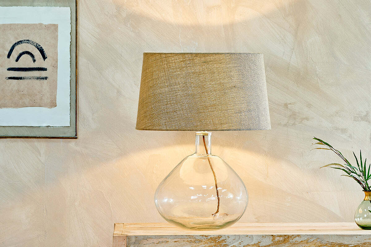 The Baba Recycled Glass Lamp - Clear - Large Wide by nkuku, featuring an elegant base and beige fabric shade, sits on a light wooden console with textured neutral walls, abstract art, and a small vase with green foliage.