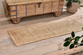 The nkuku Braided Hemp Floor Runner, a rectangular eco-friendly piece, lies on a light wooden floor by a rustic chest. Green potted plants at the edges enhance the cozy, natural look.